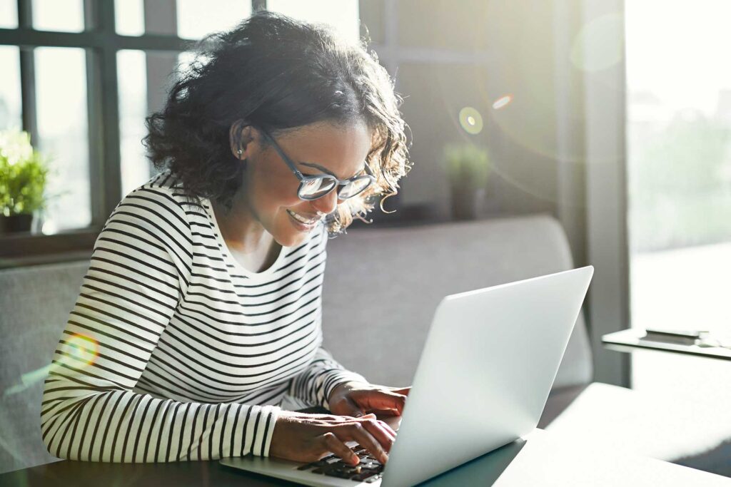 Smiling woman with glasses working on a laptop by a sunny window